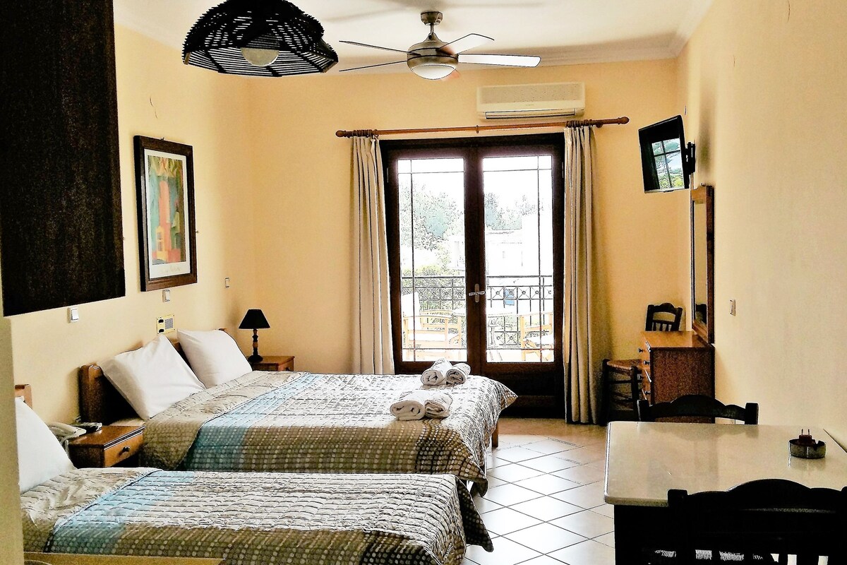 A hotel room with two beds, a ceiling fan, a window with curtains, a small table, and a TV mounted on the wall.