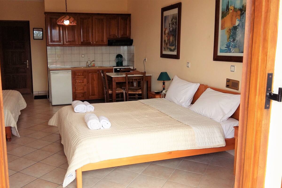 A hotel room with a bed, a desk, a chair, a small kitchenette, and a door leading to another room.