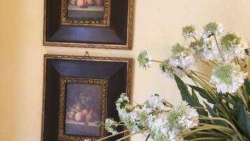Dos cuadros enmarcados con fruta en una pared, con un jarrón de flores blancas a la derecha.