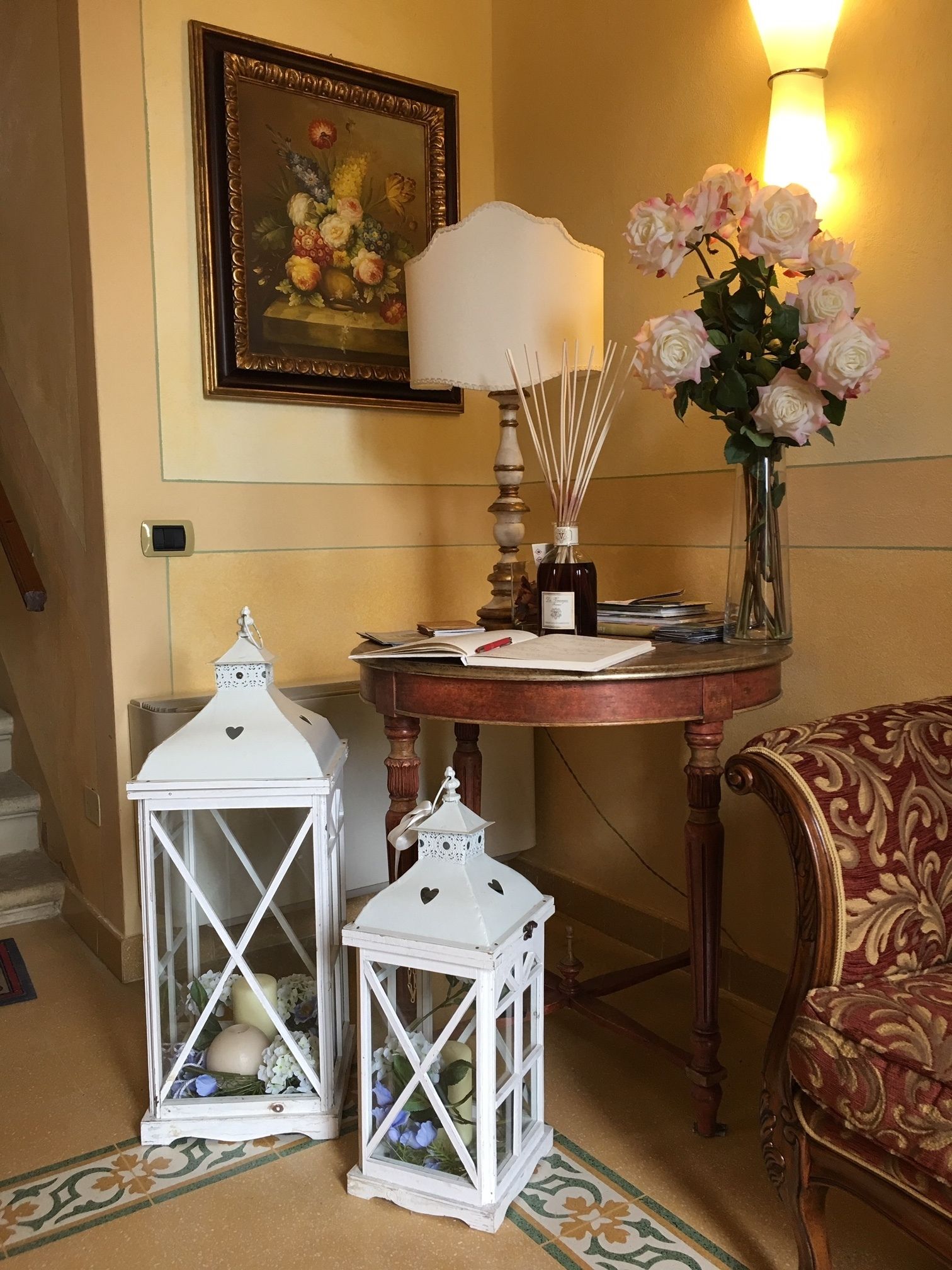 Una habitación con una mesa, dos faroles blancos, un jarrón con flores, una lámpara y un cuadro en la pared.