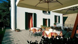 An outdoor dining area with tables and chairs, a large umbrella, and a building with green shutters. - Villa della Certosa (Gambassi Terme)