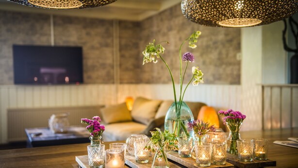Una mesa de madera con candelabros de vidrio y jarrones con flores, sobre un fondo que muestra una sala de estar con televisor y sofá.