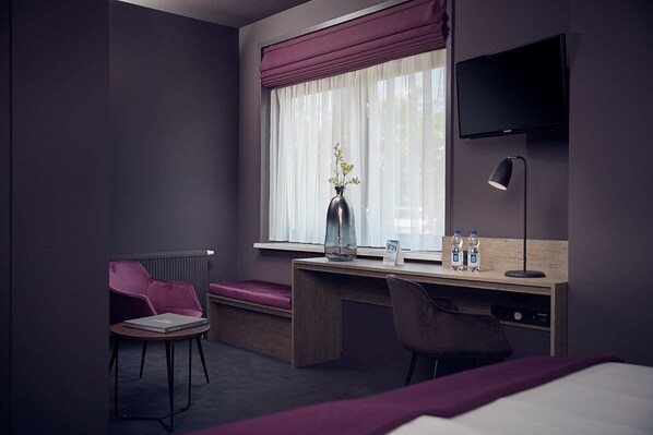 A hotel room with a wooden desk, a chair, a bed, a TV, a lamp, and a window with curtains. - Prins Hendrik Texel (Oosterend)
