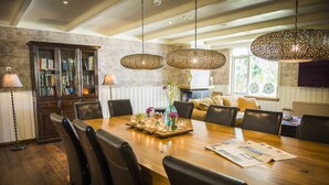 Un comedor con una mesa de madera, sillas y lámparas colgantes. Se observa una estantería y una chimenea al fondo.