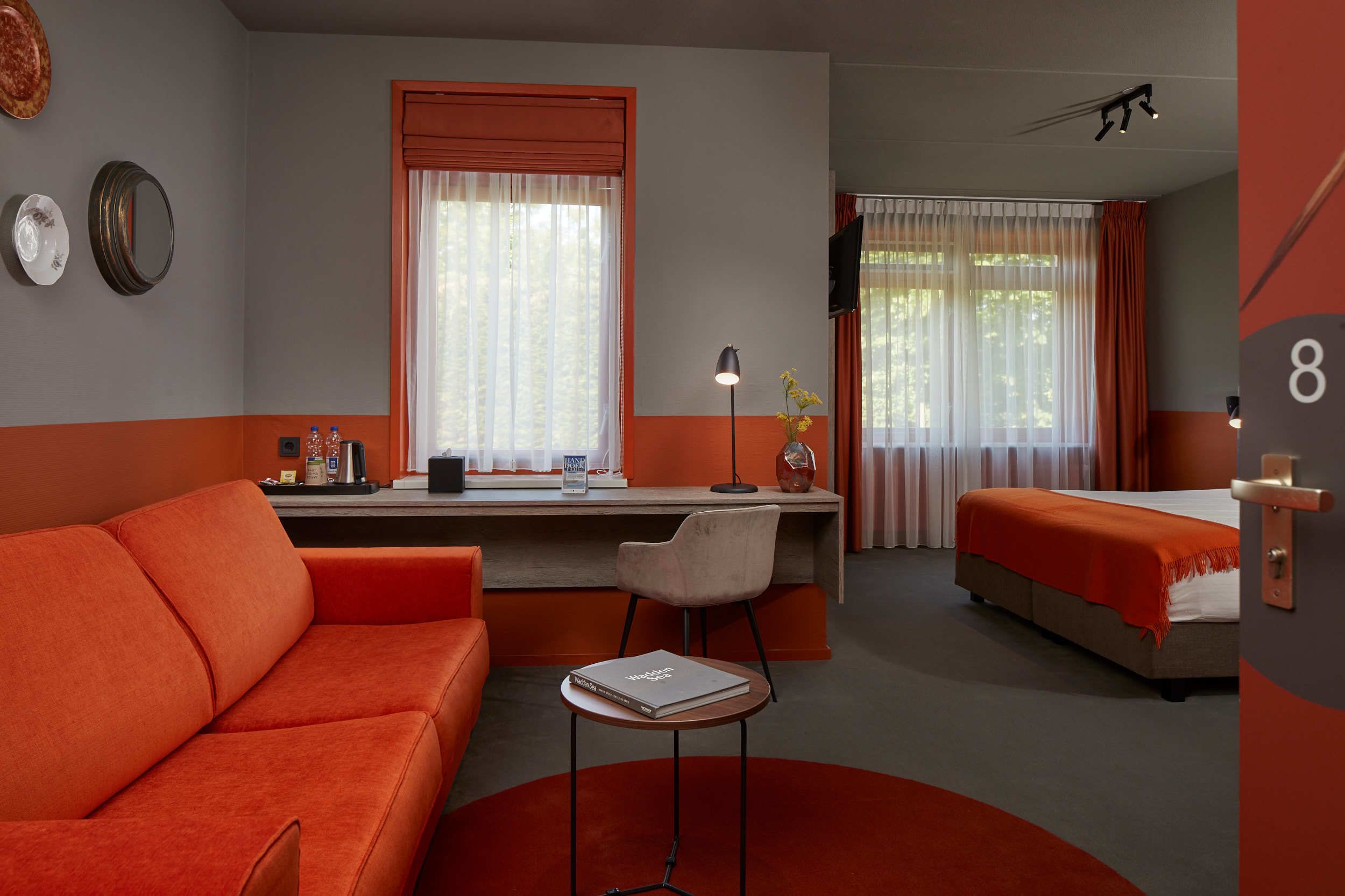 Hotellihuone, jossa on sohva, työpöytä tuolilla, sänky ja pieni pöytä, jonka päällä on kirja.