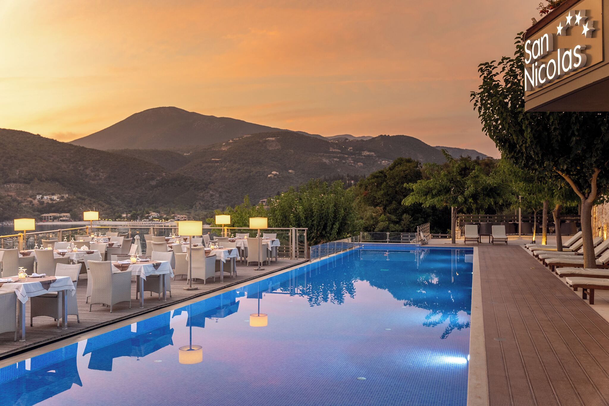 A rooftop pool area with tables and chairs, a sign reading "San Nicolas," and a view of mountains in the background.