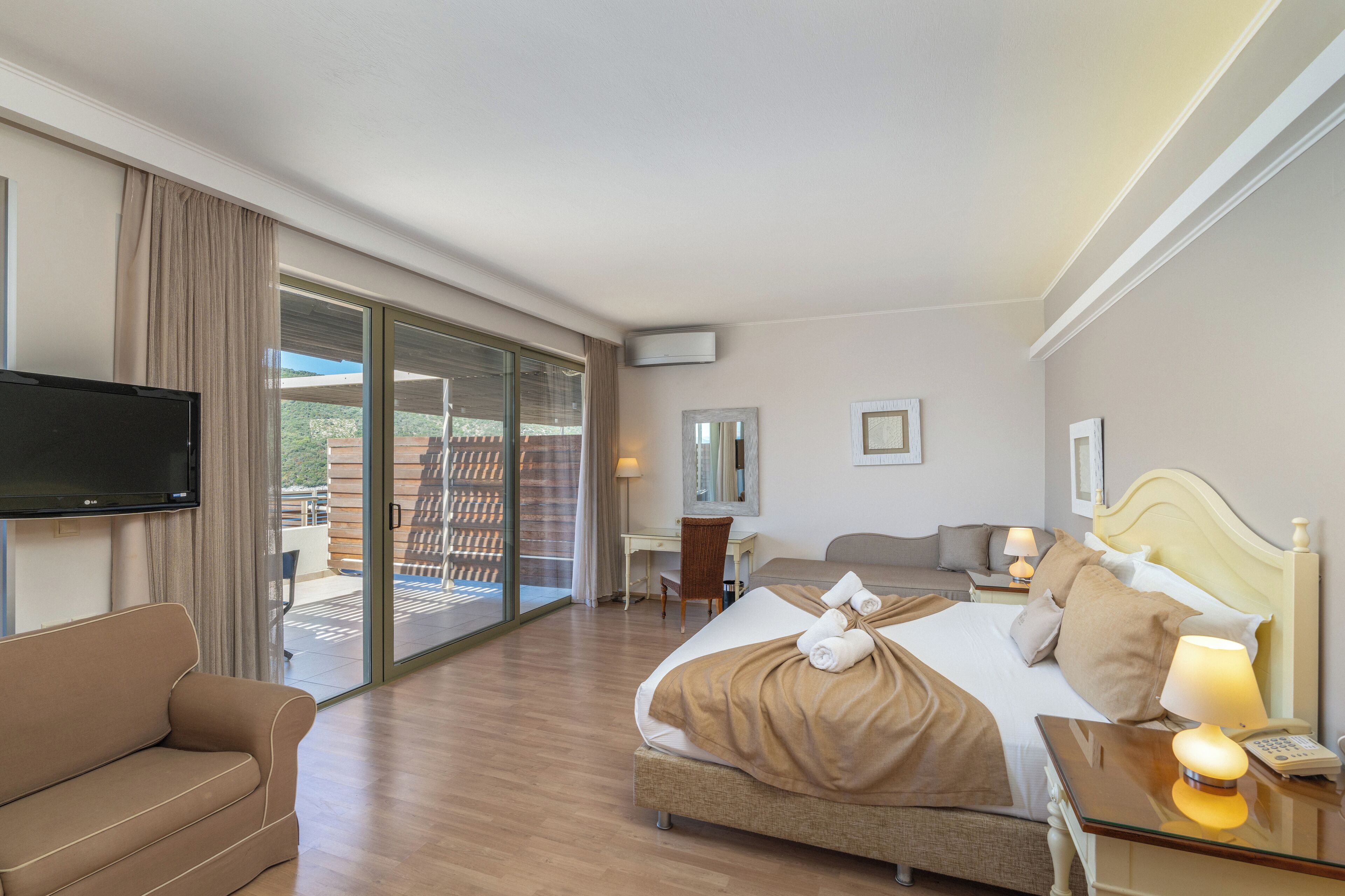 A hotel room with a bed, a sofa, a desk, a TV, and a sliding glass door leading to an outdoor area.
