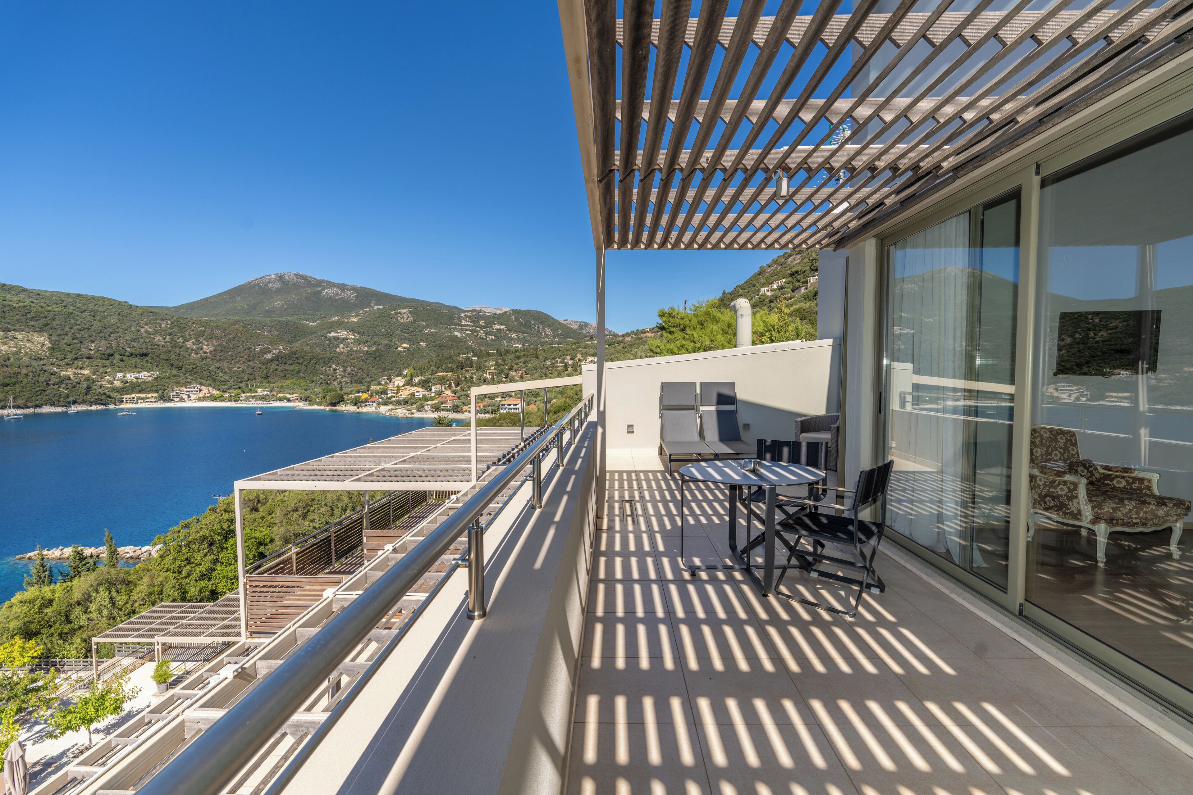 A balcony with a table and chairs, overlooking a coastal view with mountains in the background.