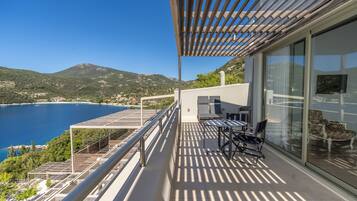 A balcony with a table and chairs, overlooking a coastal view with mountains in the background.