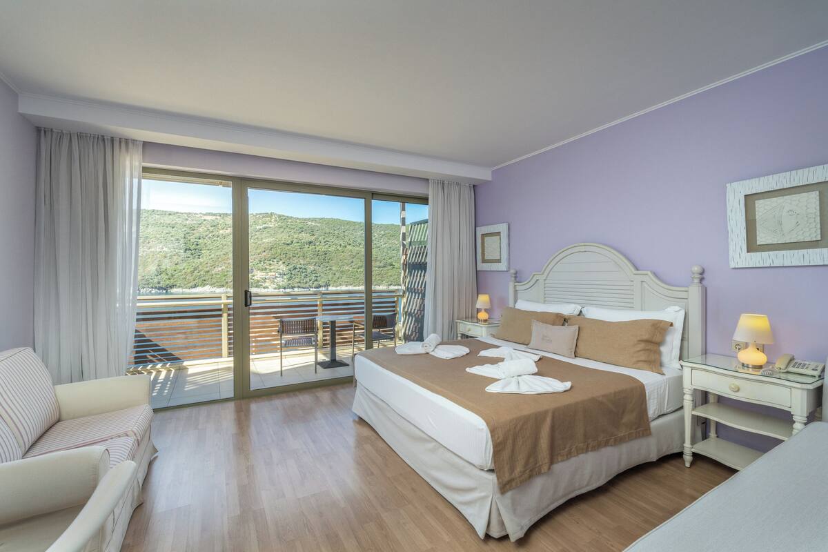 Una habitación de hotel con una cama grande, un sofá, una mesita de noche con lámpara y vistas a un balcón con barandilla.