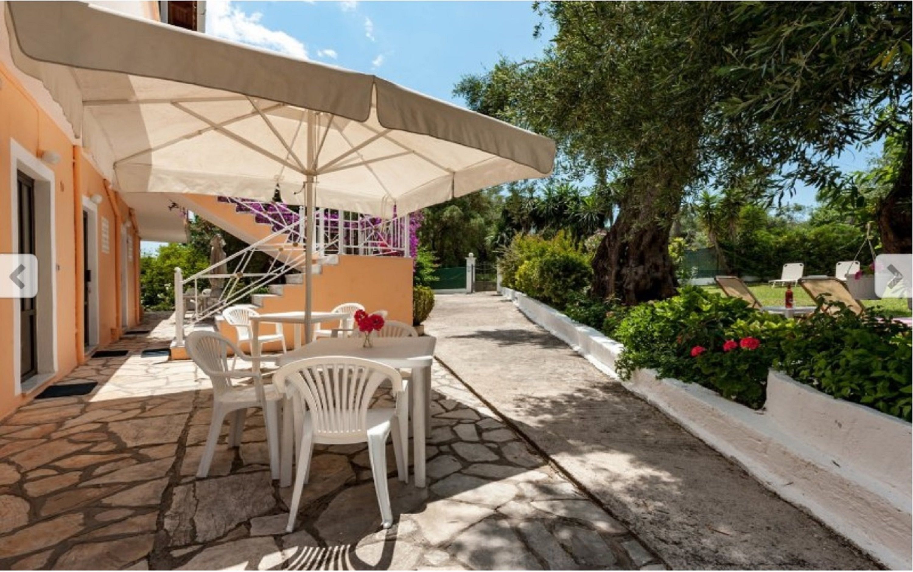 An outdoor patio with a table and chairs under a large umbrella, surrounded by stone flooring and greenery.