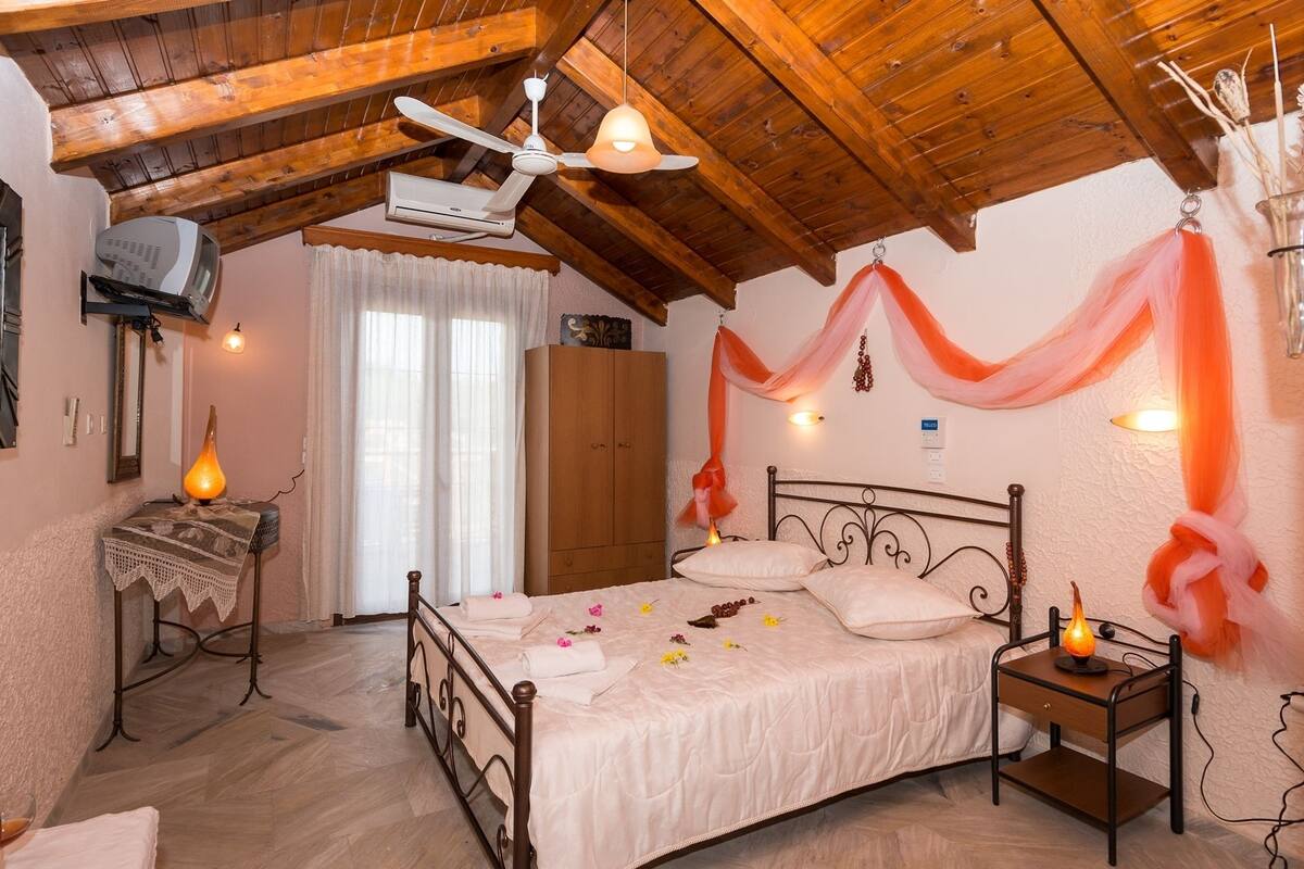 A bedroom with a wooden ceiling, a bed with white linens, a bedside table with a lamp, and a wall-mounted air conditioner.