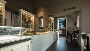 A modern bar area with a long counter, glass display case with fruits, and a view of trees through the window. - Villa Avellino (Pozzuoli)
