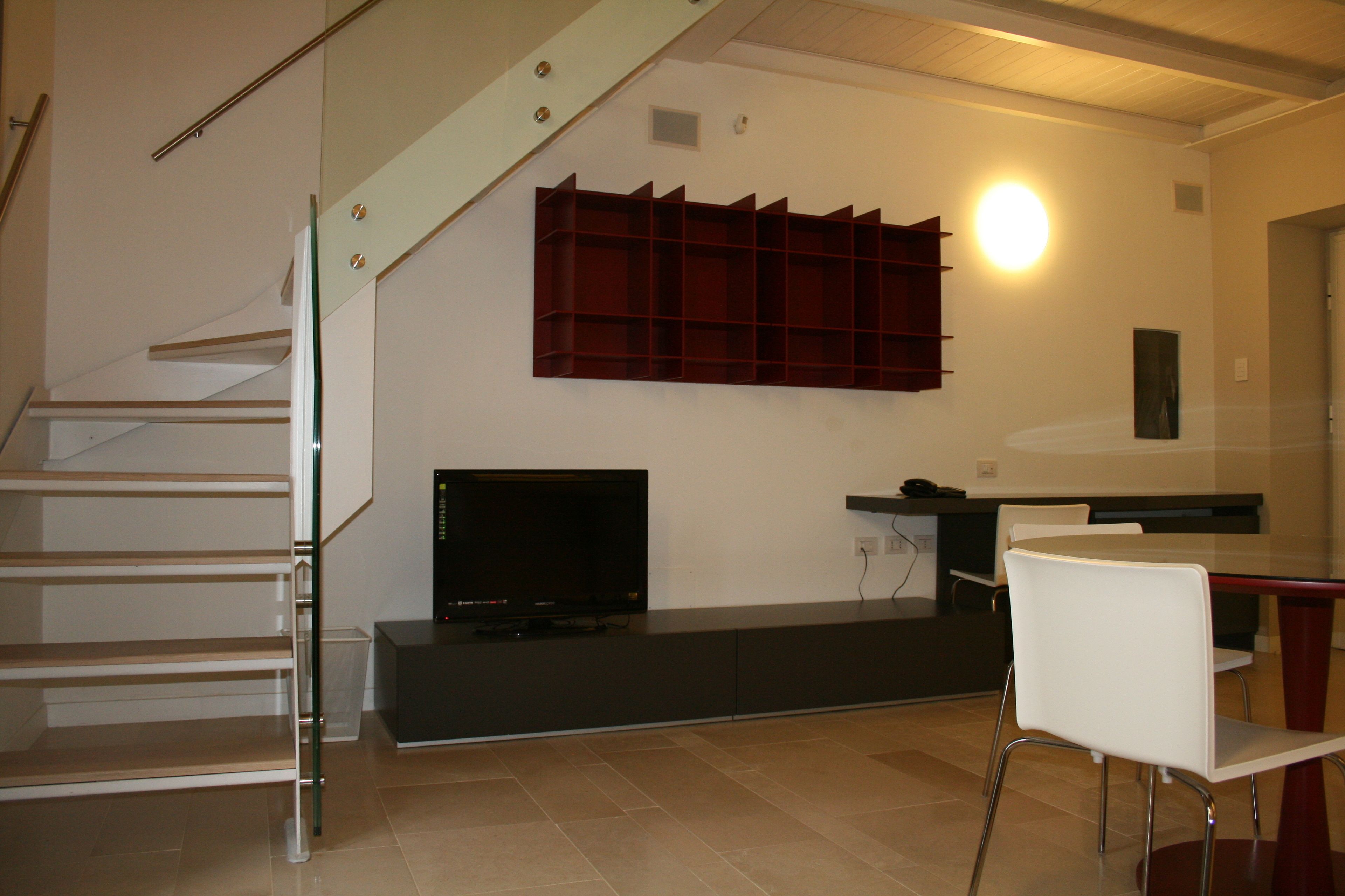 A modern living room with a staircase, a flat-screen TV, a dining table with chairs, and a built-in shelving unit.