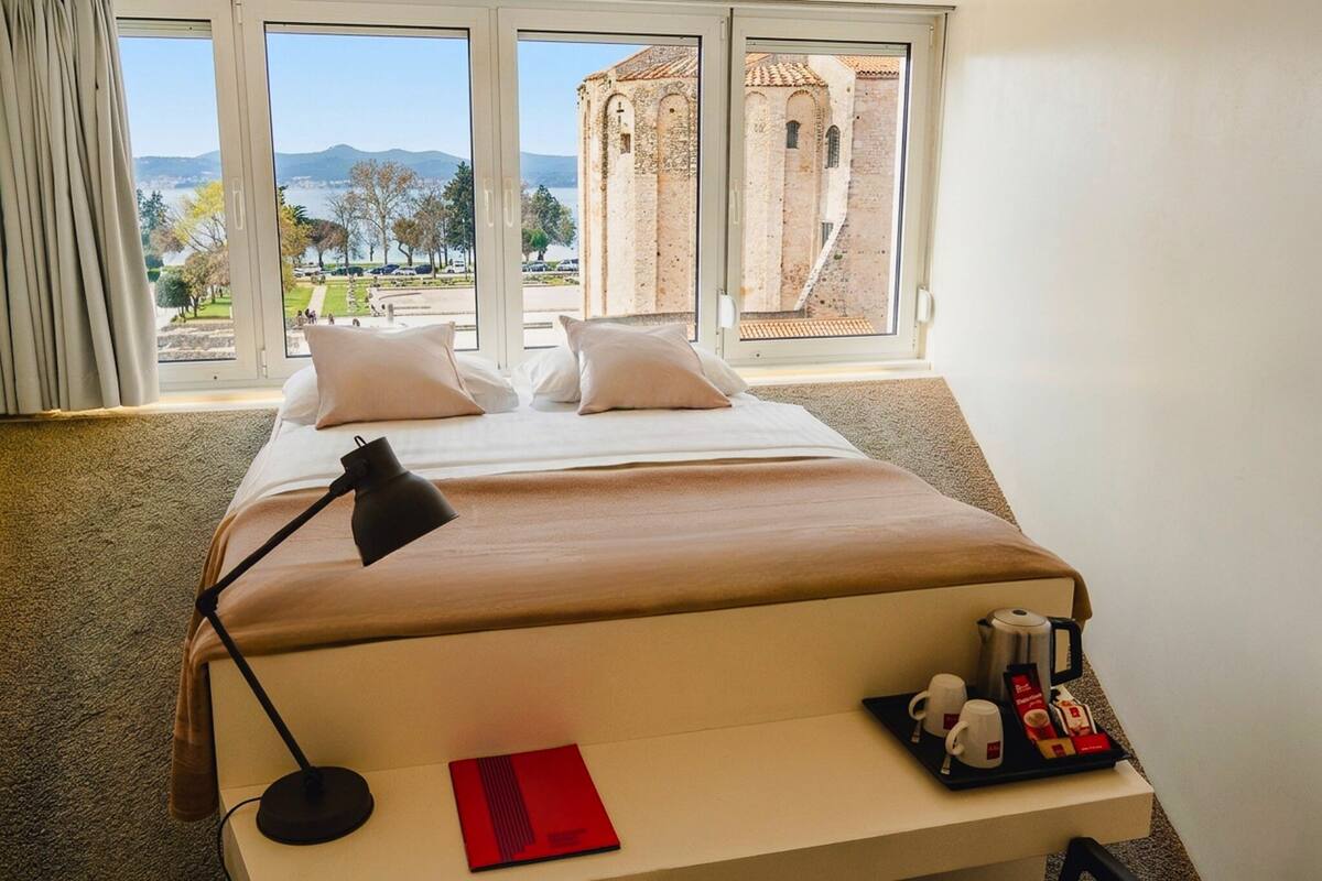 A hotel room with a bed, a desk with a lamp, a tray with coffee and tea, and a view of a historic building and a park.