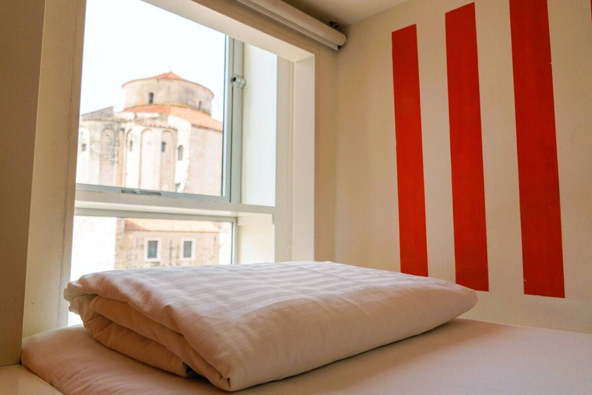 A room with a bed, a window, and a view of a building outside.