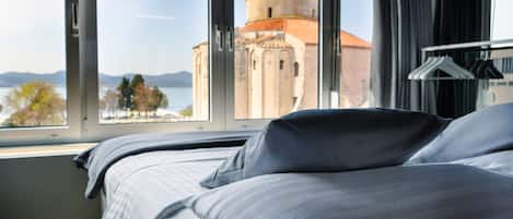 A hotel room with a bed, a window with a view of a historic building, and a coat rack.