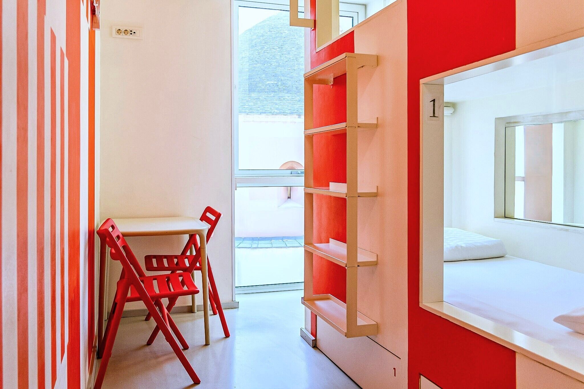 Une chambre d’hôtel moderne avec un lit blanc, un mur rayé de rouge et de blanc, une chaise rouge et une fenêtre donnant sur un beau paysage.