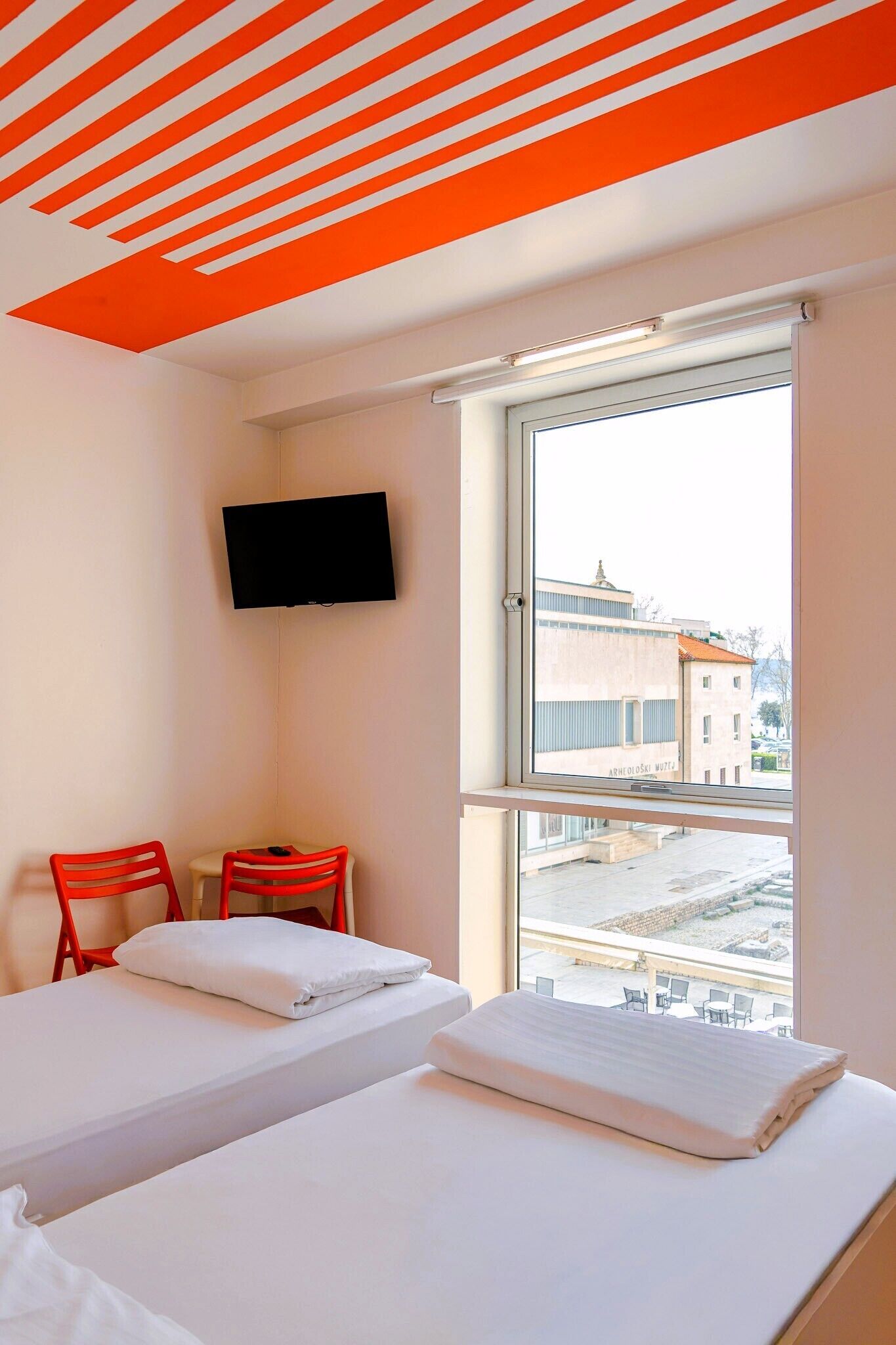 A hotel room with two beds, a TV mounted on the wall, and a window with a view of buildings.