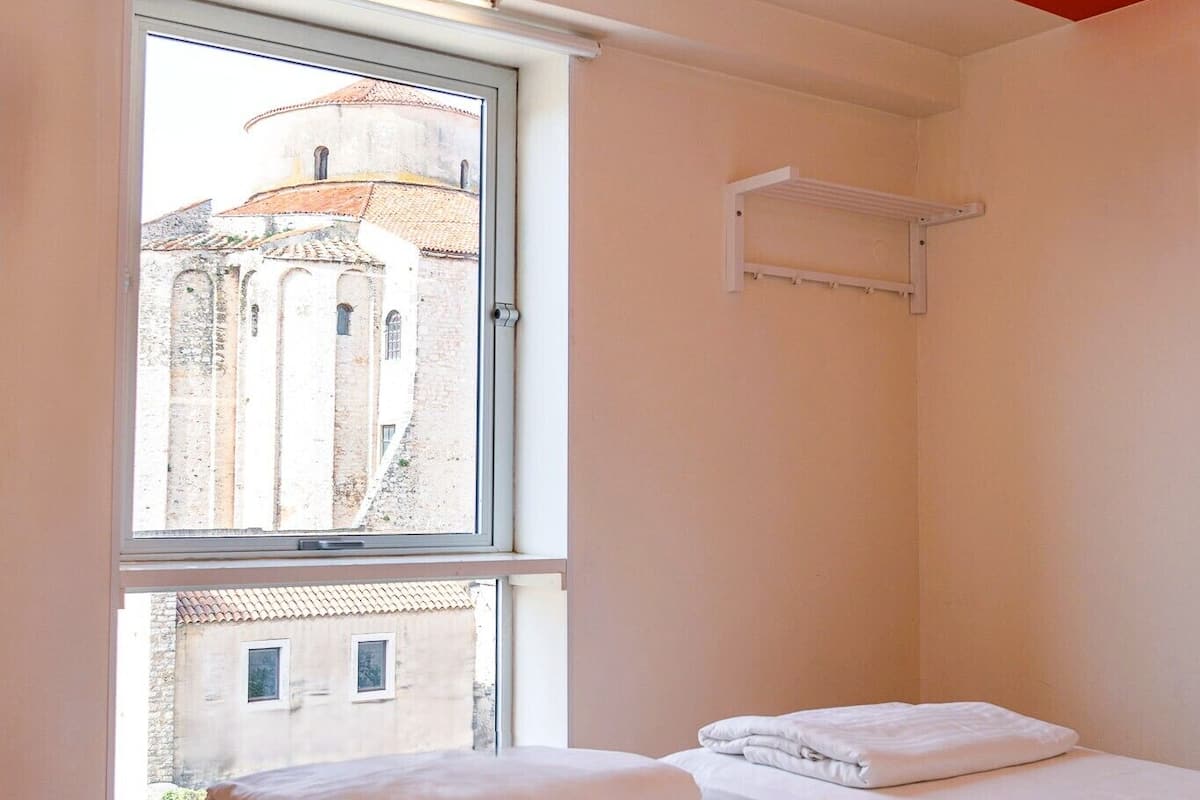 A room with two beds, a window with a view of a building, and a ceiling with orange stripes.