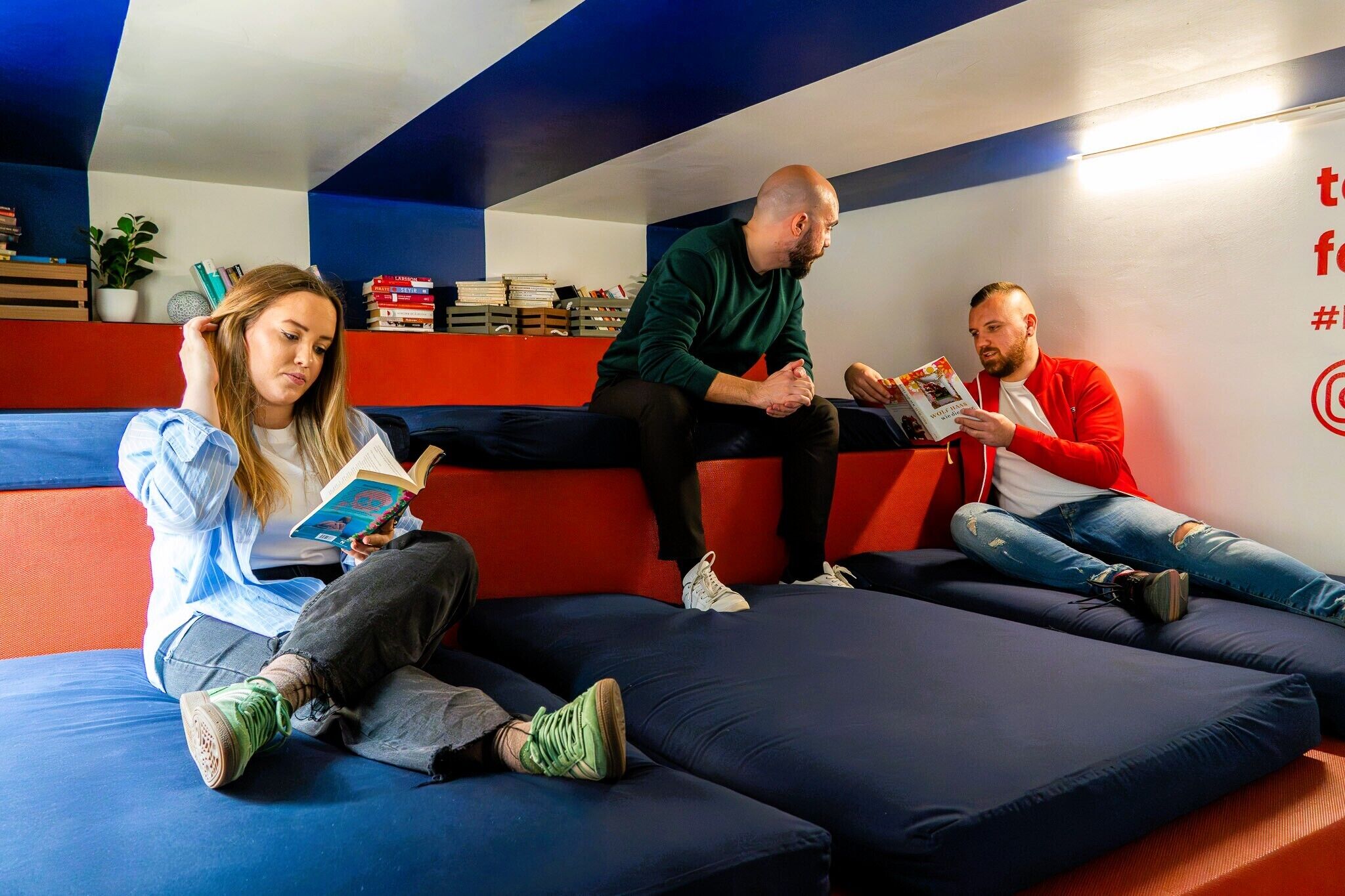 Drei Personen sitzen auf einem Sofa in einem Raum mit roten und blauen Wänden und lesen Bücher.