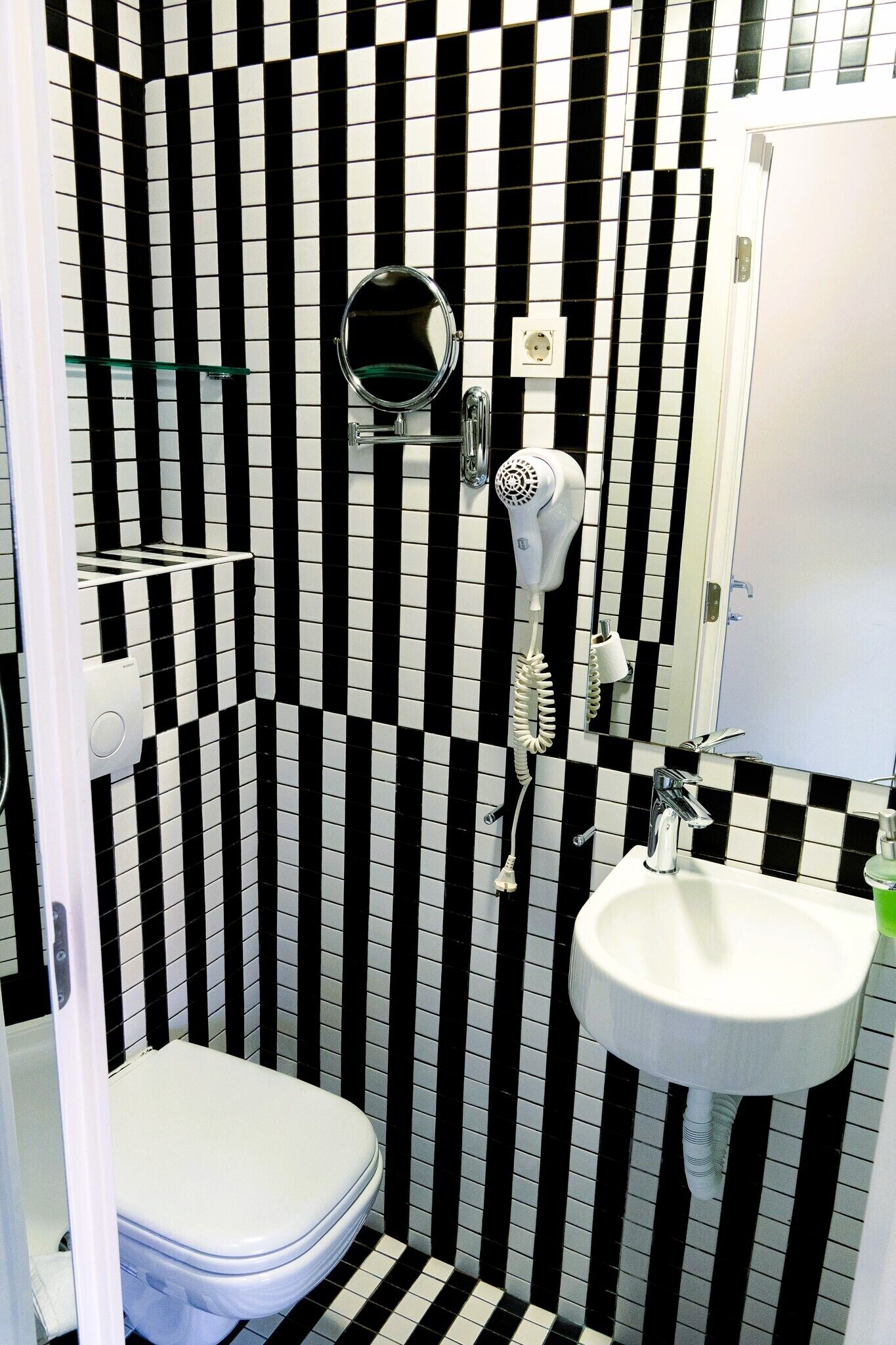 Un bany amb una paret de rajoles blanquejades i negres a ratlles, un vàter blanc, un lavabo i un assecador de cabells fixat a la paret.