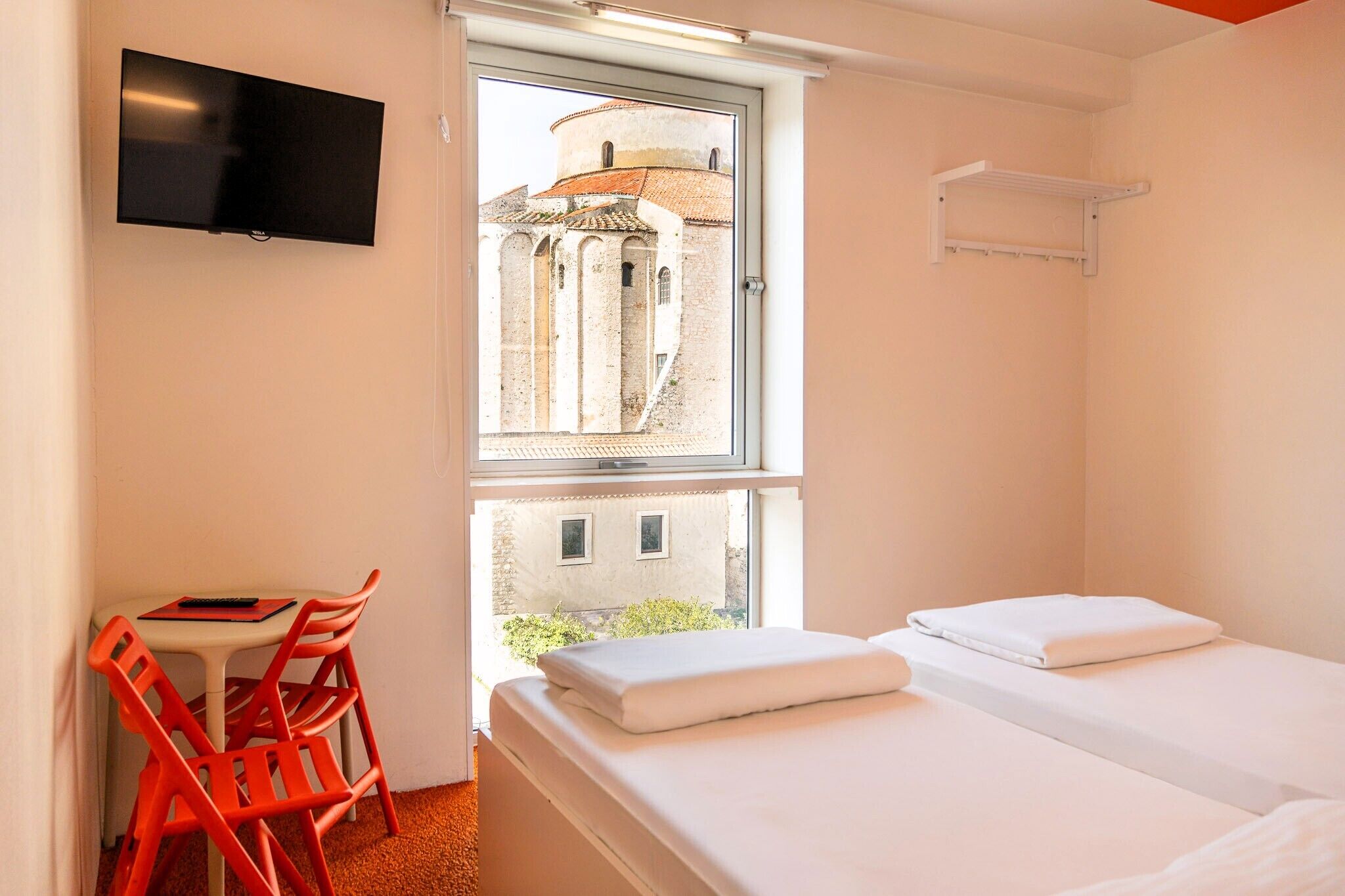 A room with a bed, a chair, a TV, and a window with a view of a building.