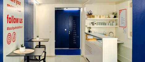 Une cuisine moderne avec un mur d’accent bleu, un comptoir blanc et un petit îlot central agrémenté d’une plante. On y trouve deux tables avec des chaises, un évier et une porte dotée d’un clavier numérique.