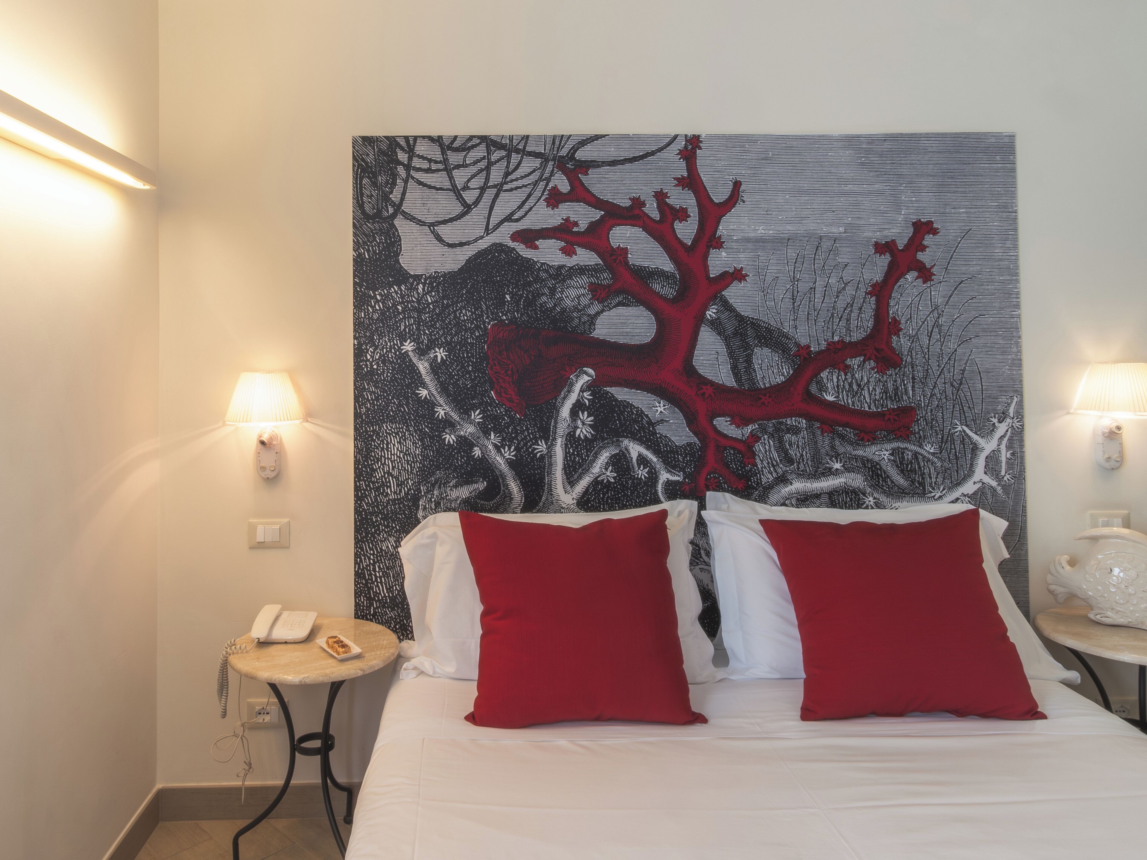 Ein Hotelzimmer mit einem Bett, zwei roten Kissen, einem Nachttisch mit Lampe und einer Wandteppich.