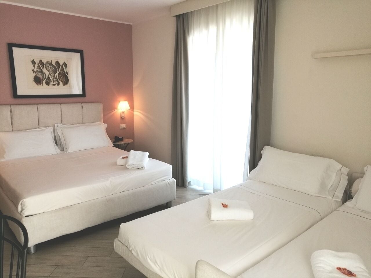 Een hotelkamer met twee bedden, een nachtkastje met een lamp, een kunstwerk in een lijst aan de muur, en een raam met gordijnen.