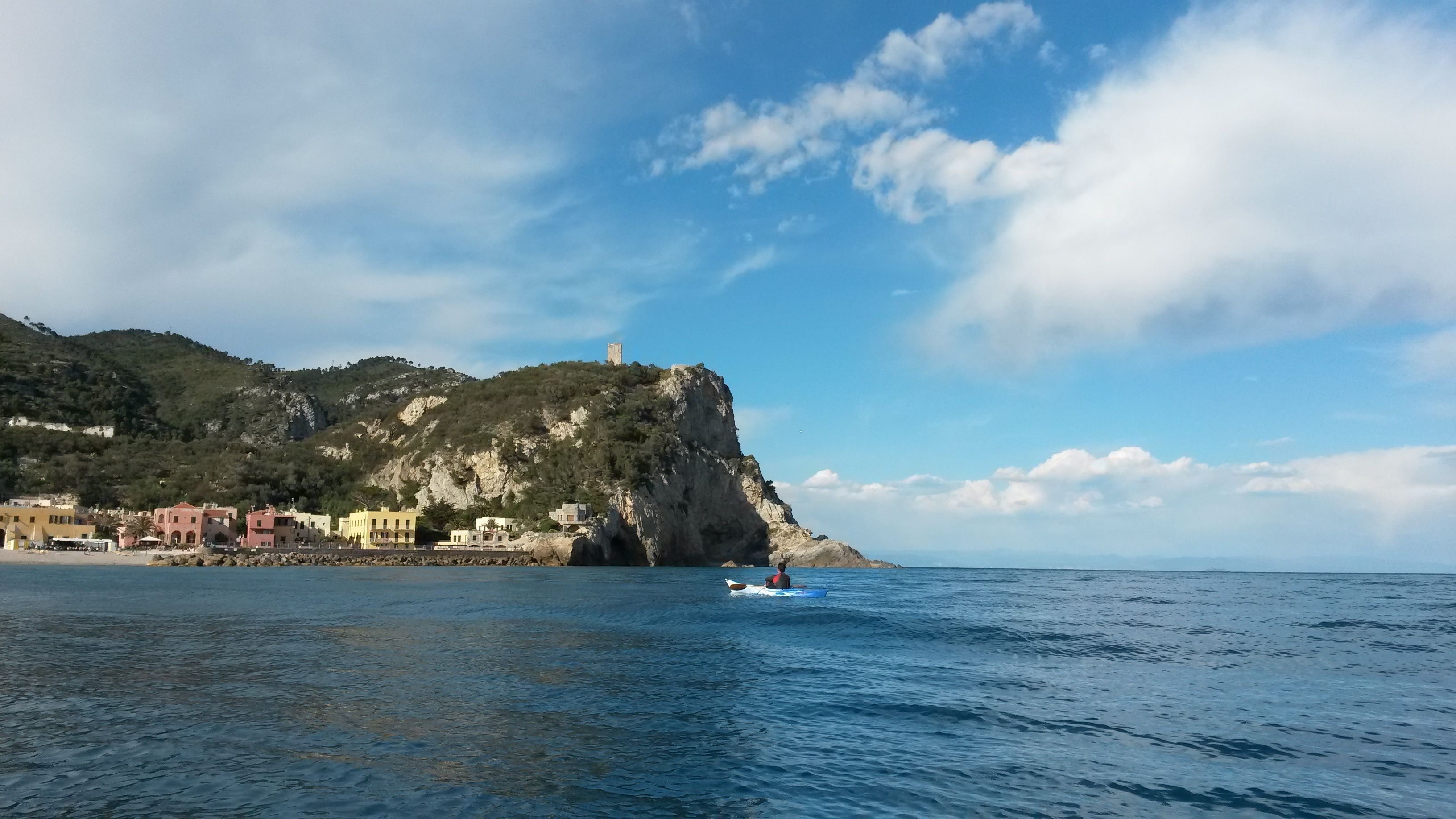 A coastal town with buildings along the shore, a prominent cliff with a tower, and calm sea waters.