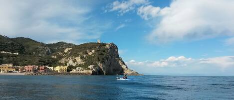 A coastal town with buildings along the shore, a prominent cliff with a tower, and calm sea waters.