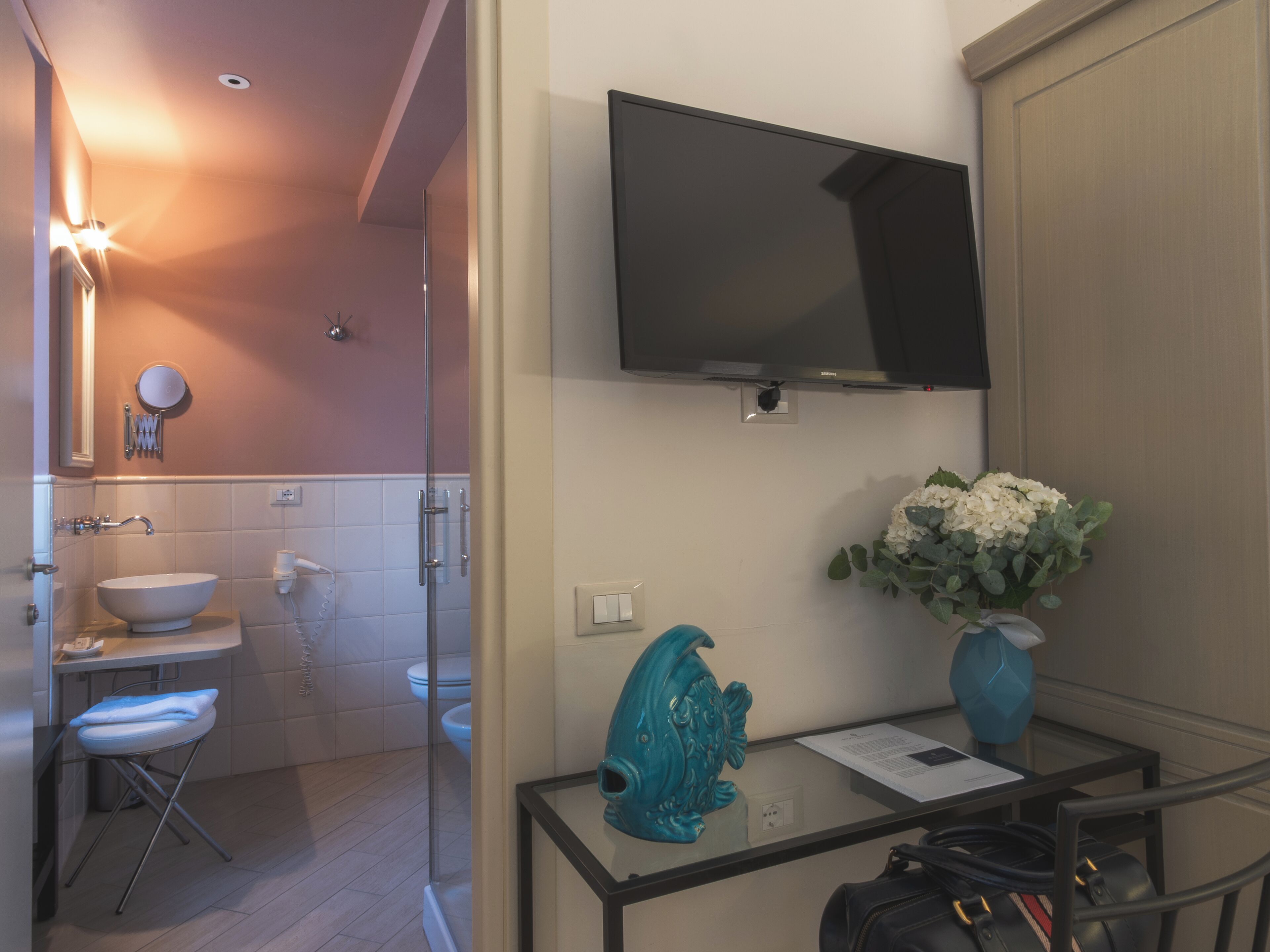 Un baño con lavabo, inodoro y ducha. Hay un televisor montado en la pared, un florero con flores, y una mesa con una bolsa y papeles.