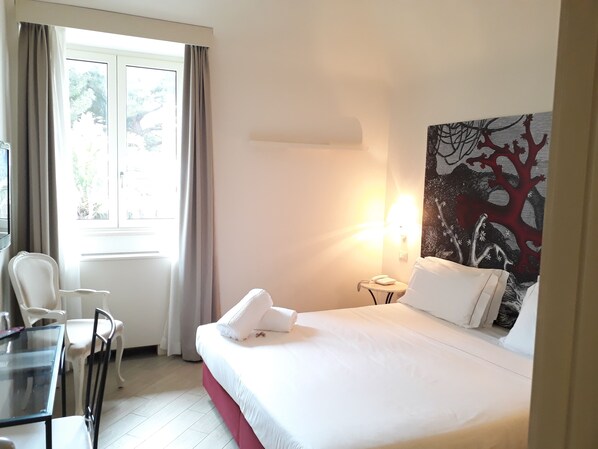 A hotel room with a bed, a desk, chairs, a window with curtains, and a wall with a tree painting. - San Pietro Palace Hotel (Finale Ligure)