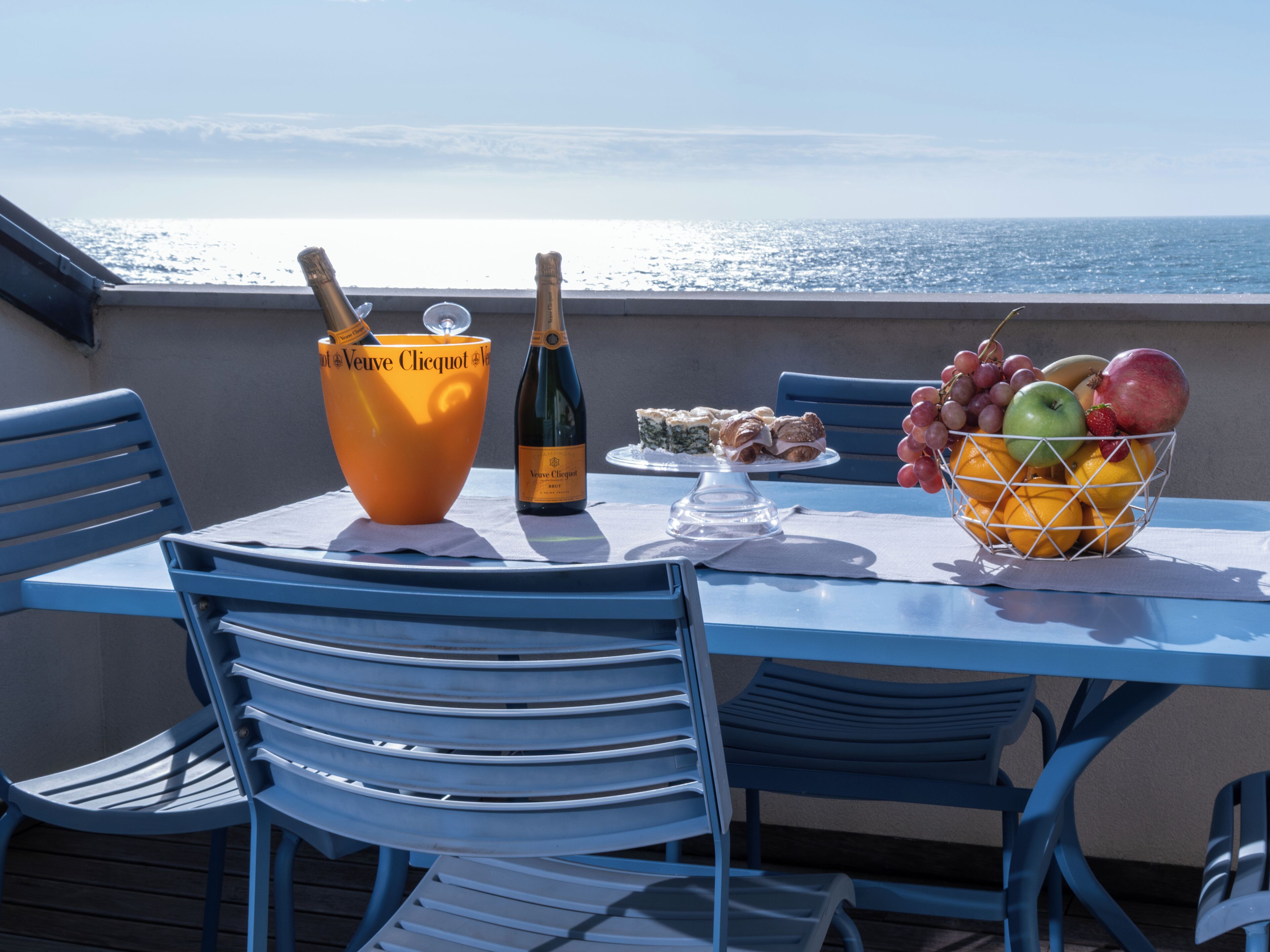 A table with a bottle of Veuve Clicquot champagne, a bowl of fruit, and a dessert plate on a balcony overlooking the sea.
