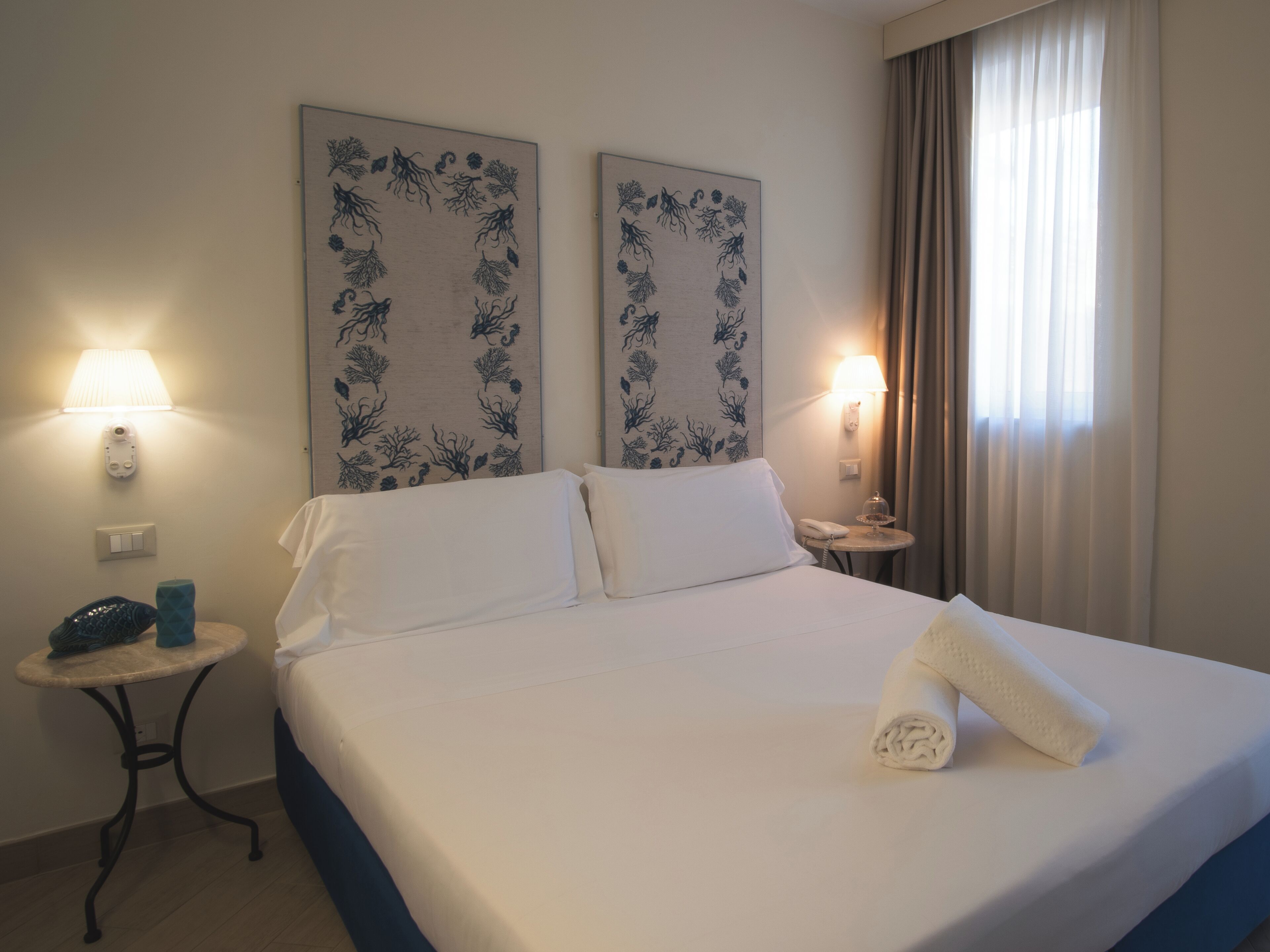 A hotel room with a bed, two wall-mounted lamps, a bedside table with a phone, and a window with curtains.