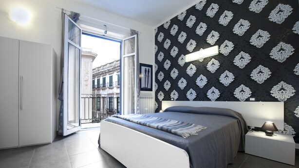 A modern bedroom with a large bed, a nightstand, a window with an open door, and a patterned wall. - Gran Maestro B&B (Palermo)