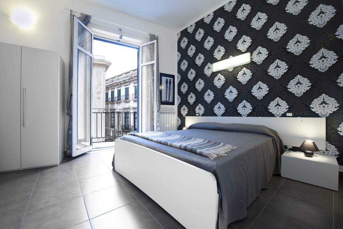 A modern bedroom with a large bed, a nightstand, a window with an open door, and a patterned wall.