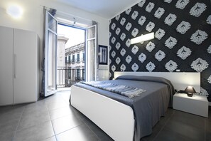 A modern bedroom with a large bed, a nightstand, a window with an open door, and a patterned wall. - Gran Maestro B&B (Palermo)