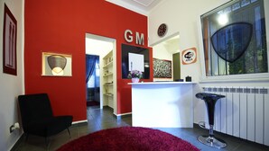 A reception area with a red wall, a black chair, a white reception desk, a black bar stool, and a framed certificate on the wall. - Gran Maestro B&B (Palermo)