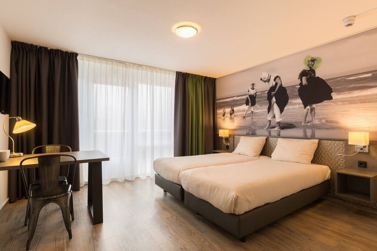 Quarto de hotel com cama grande, duas mesas de cabeceira, escrivaninha com cadeira e uma janela ampla com cortinas.