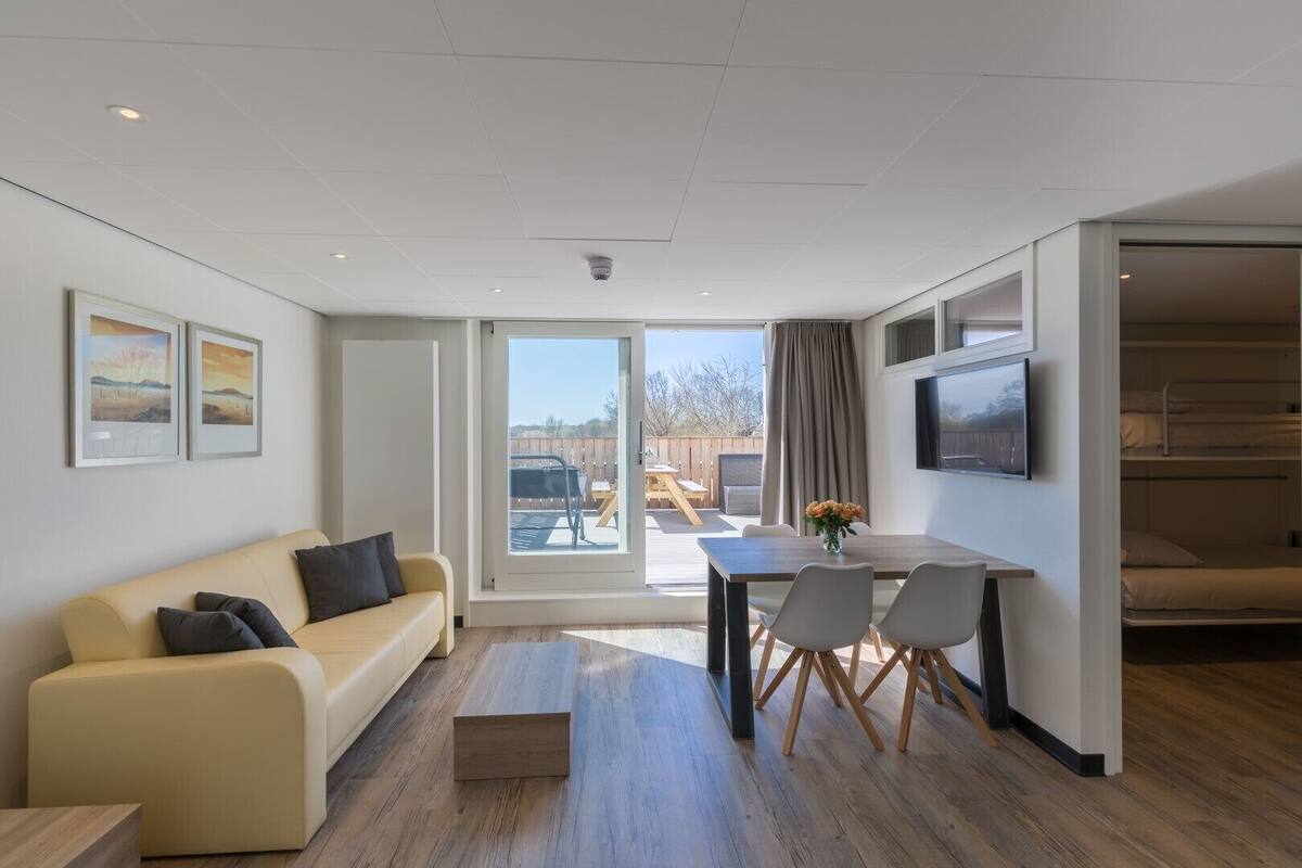 Quarto de hotel moderno com sofá, mesa de jantar com cadeiras, televisão na parede e uma janela grande com vista para o exterior.