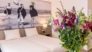 Une chambre d’hôtel avec un lit, une table de chevet, une lampe fixée au mur et un grand tableau représentant des danseurs.