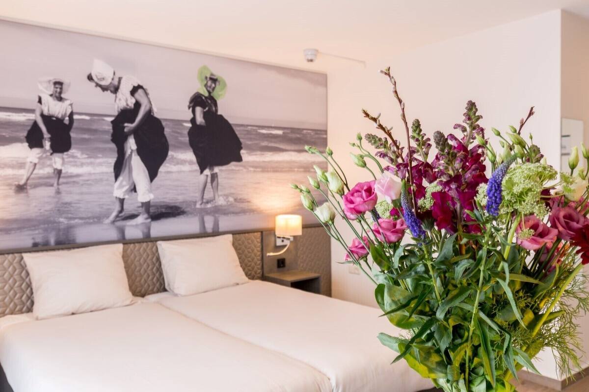 Quarto de hotel com uma cama, mesa de cabeceira, luminária na parede e uma pintura grande de dançarinos.