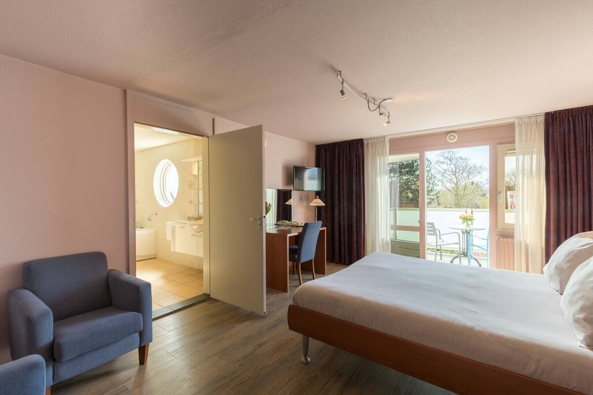 Quarto de hotel com cama, escrivaninha com cadeira, televisão e banheiro visível por uma porta aberta.