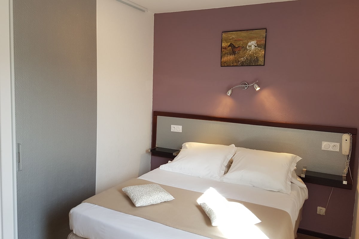 Una cama bien hecha con sábanas blancas y un cojín estampado, un aplique de pared y un cuadro de caballos.