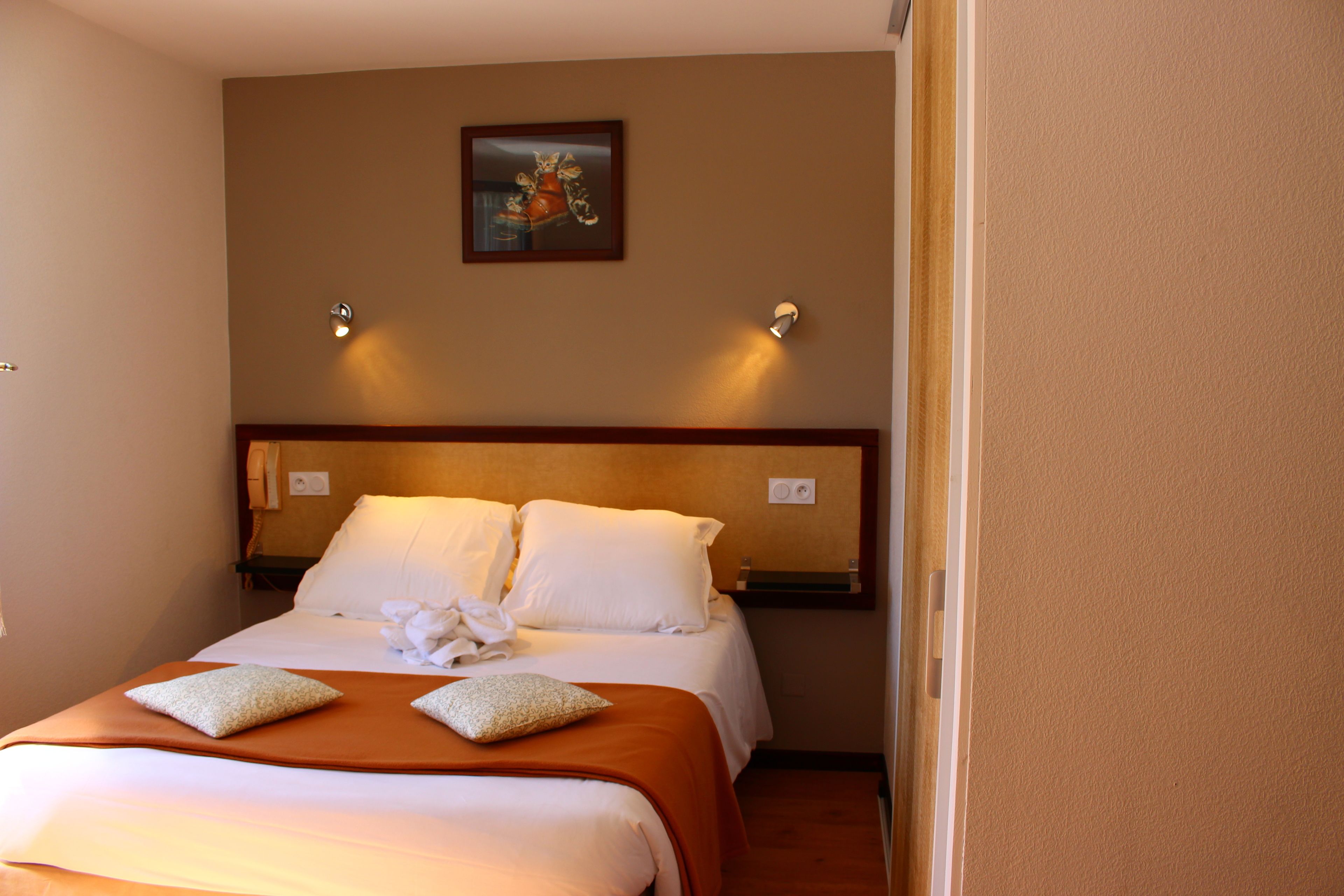 Une chambre d’hôtel avec un lit, des oreillers, une couverture, un tableau au mur et deux lampes fixées au mur.
