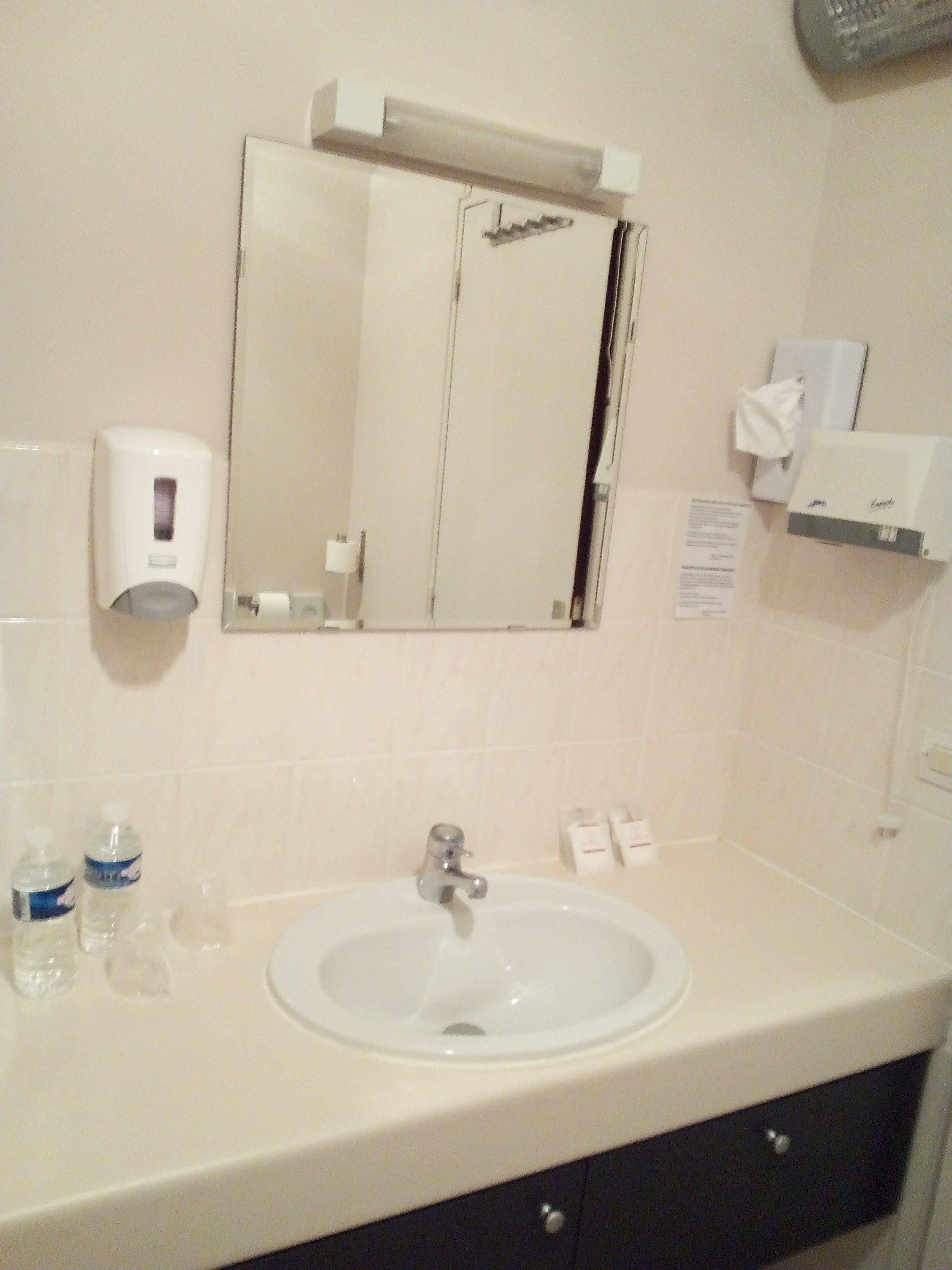 Un baño con lavamanos, espejo, dispensador de jabón y dispensador de toallas de mano.
