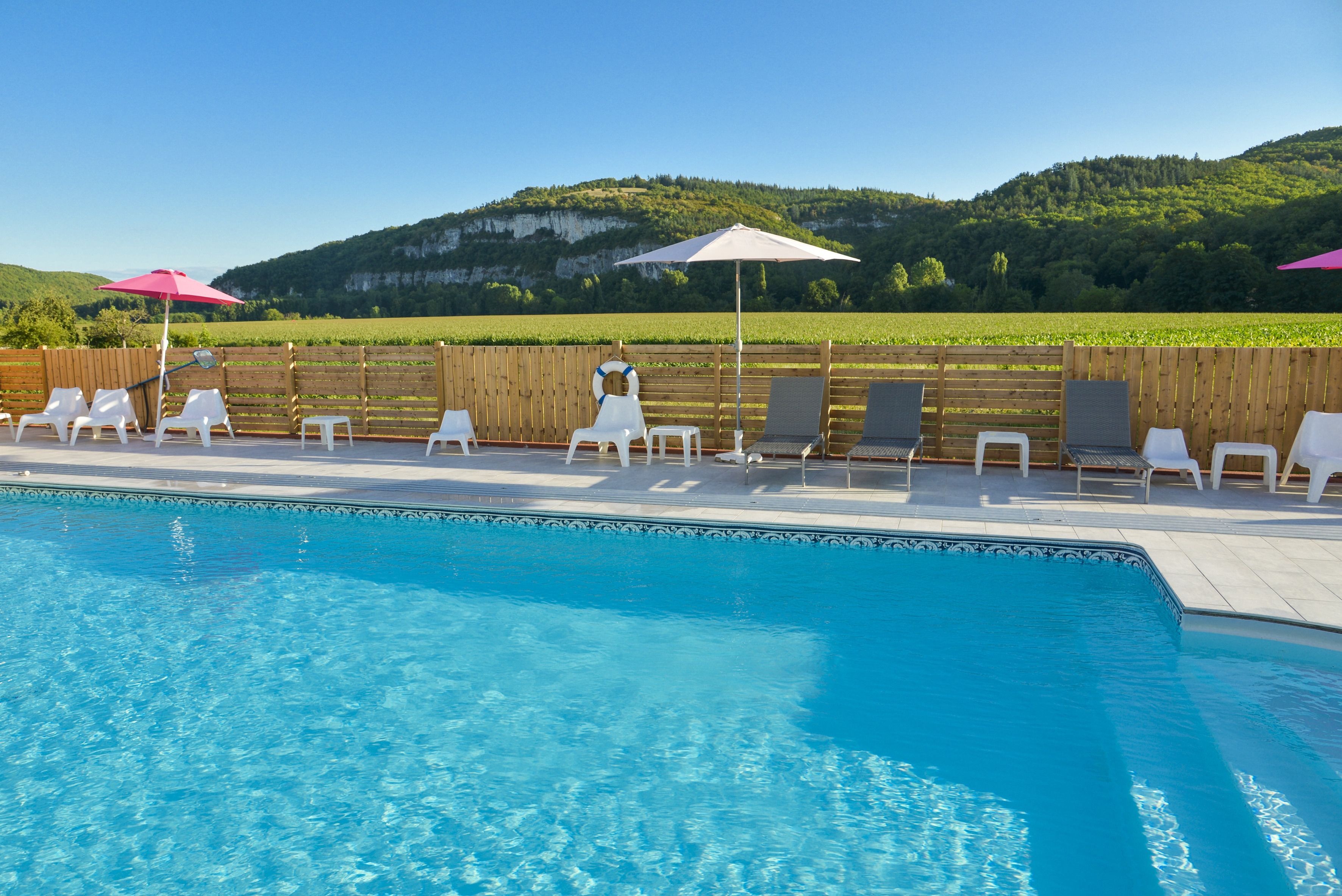 Una piscina con sillas reclinables y sombrillas blancas, rodeada por una cerca de madera y vistas a colinas verdes.