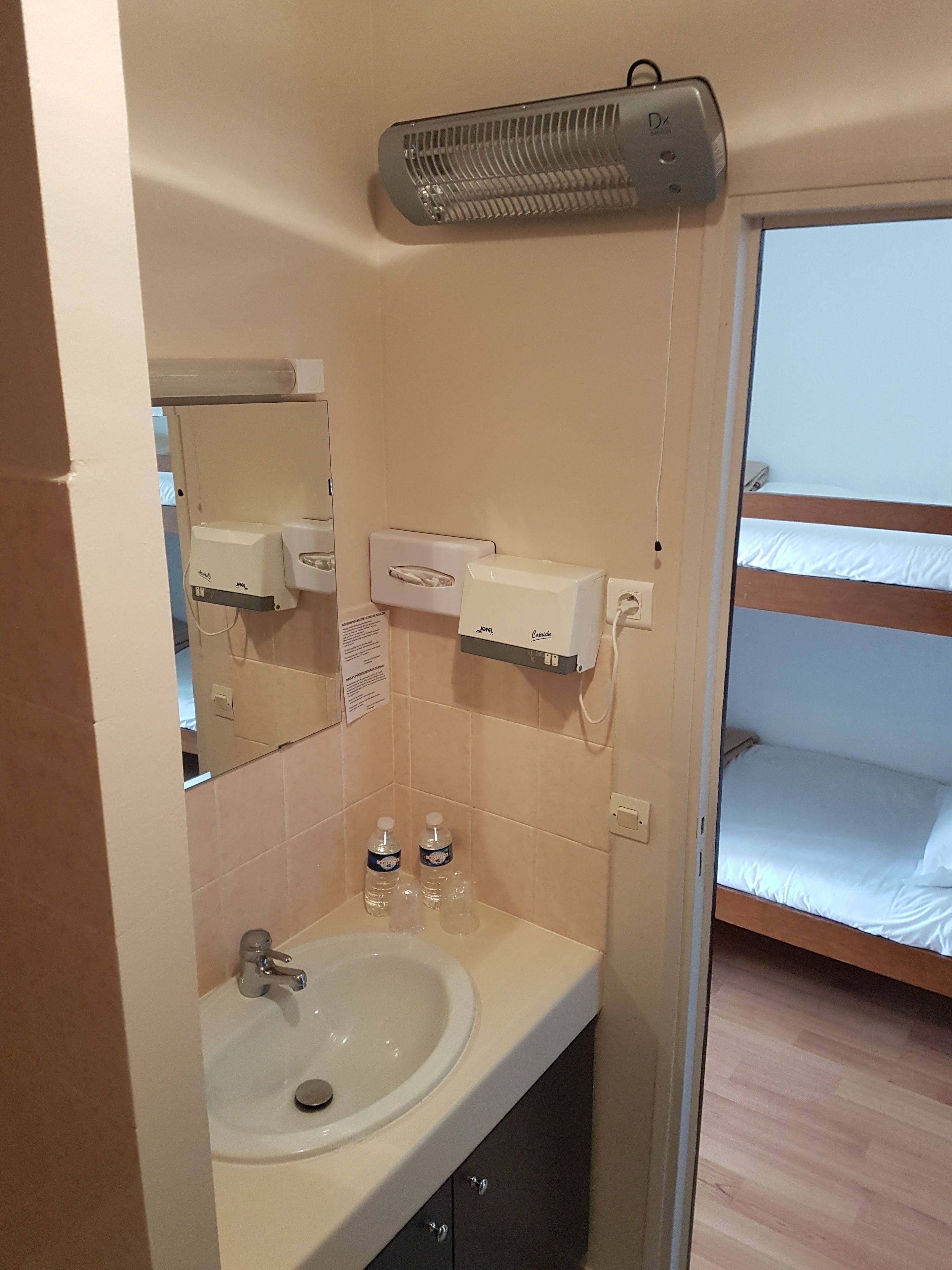 A bathroom with a sink, mirror, hand dryer, and a heater. There is a bed with a wooden headboard and white bedding.